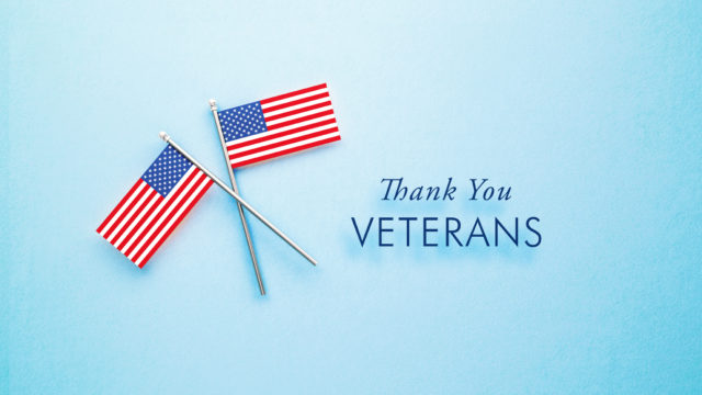 Veteran's Day Message Sitting Next To Tiny American Flag Pair On Blue Background